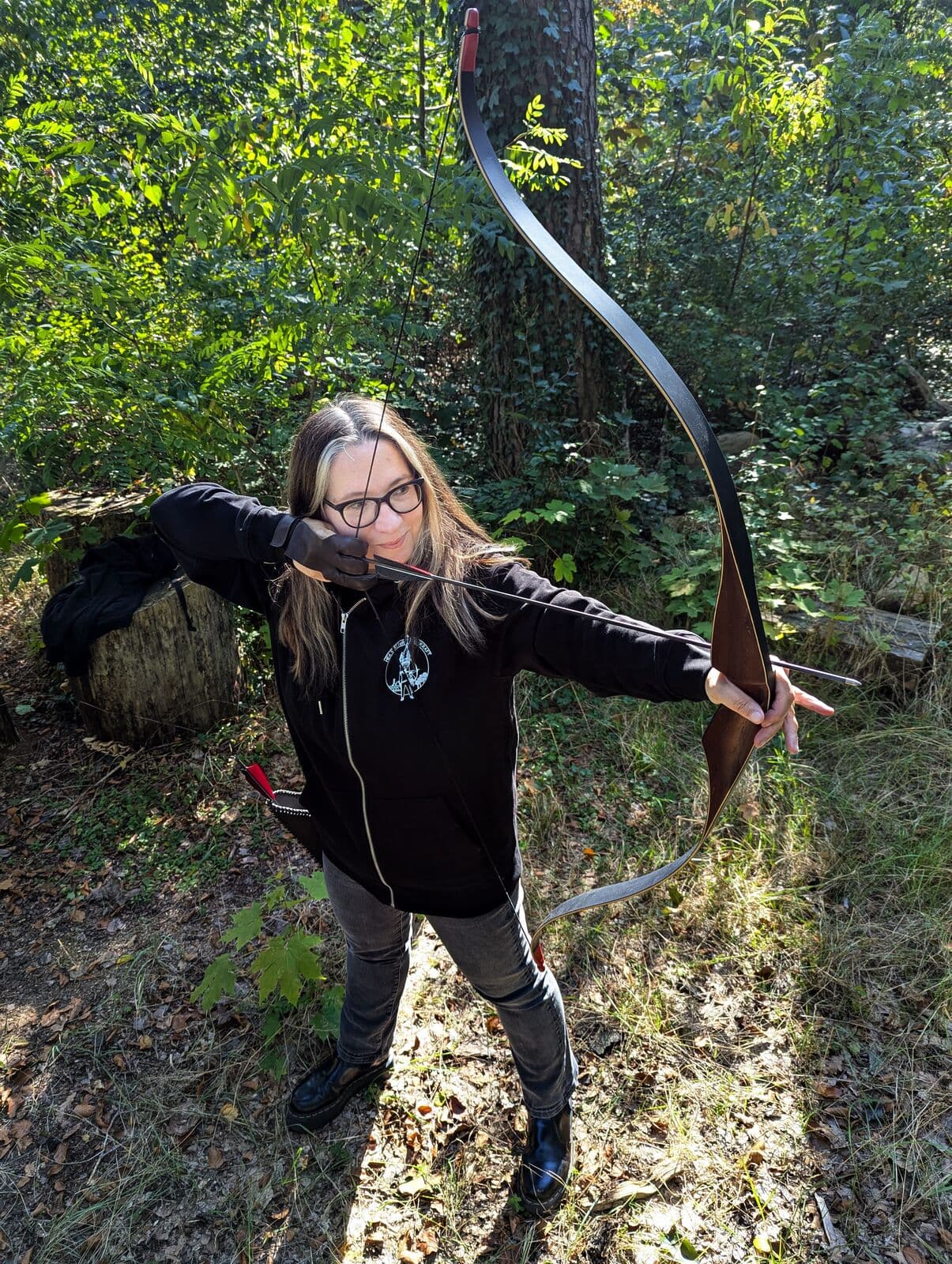 Bogenschützin im Wald spannt einen Langbogen und lächelt in die Kamera