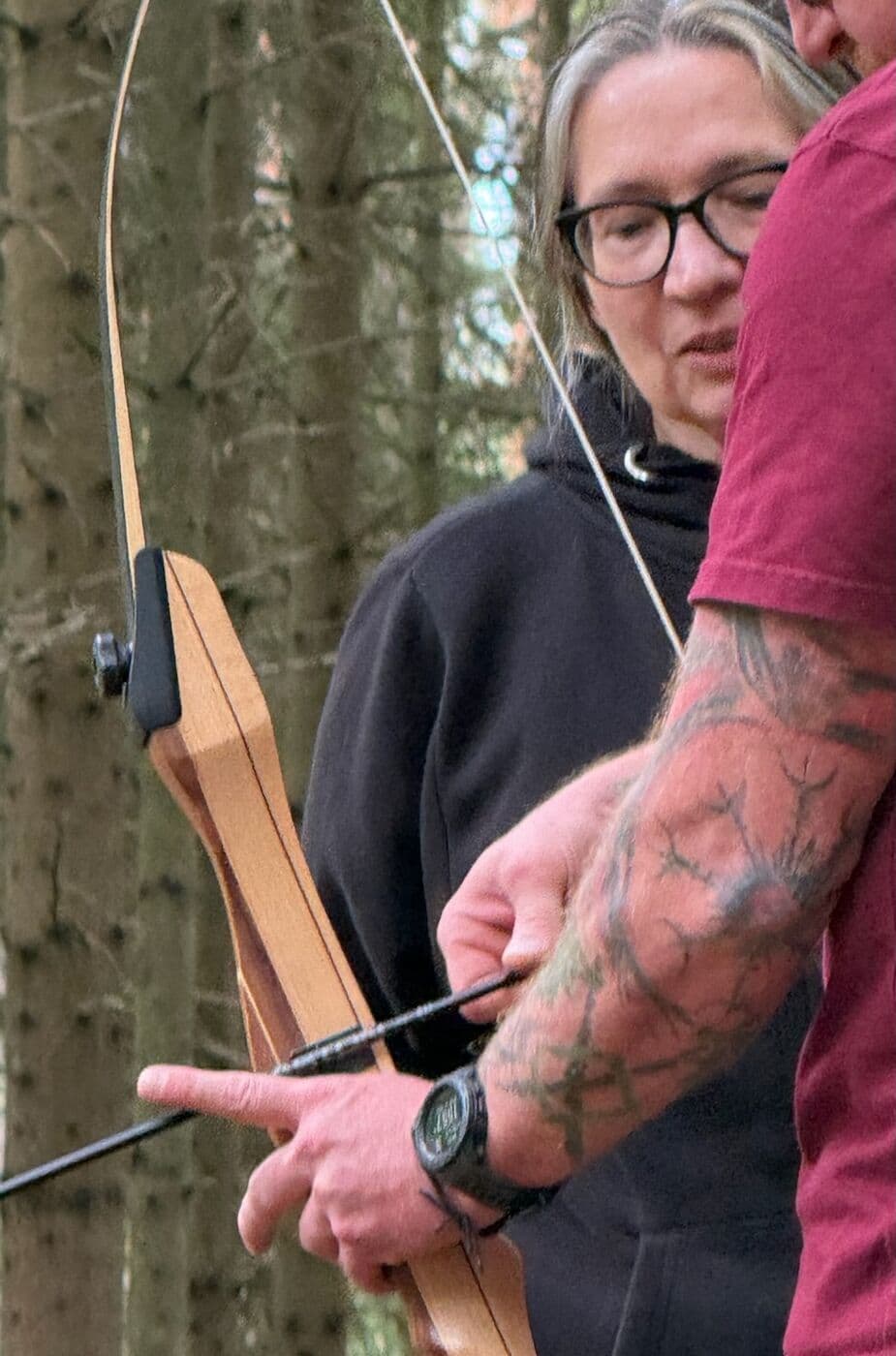 Ein Mann mit tätowiertem Unterarm zeigt einer Frau mit Brille, wie der Pfeil am Holzbogen angelegt wird